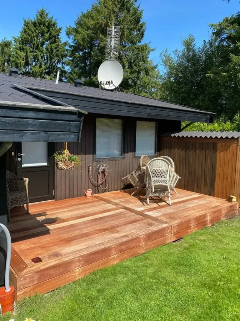 Træterrasse med havemøbler foran hus med sort tag og grøn have.